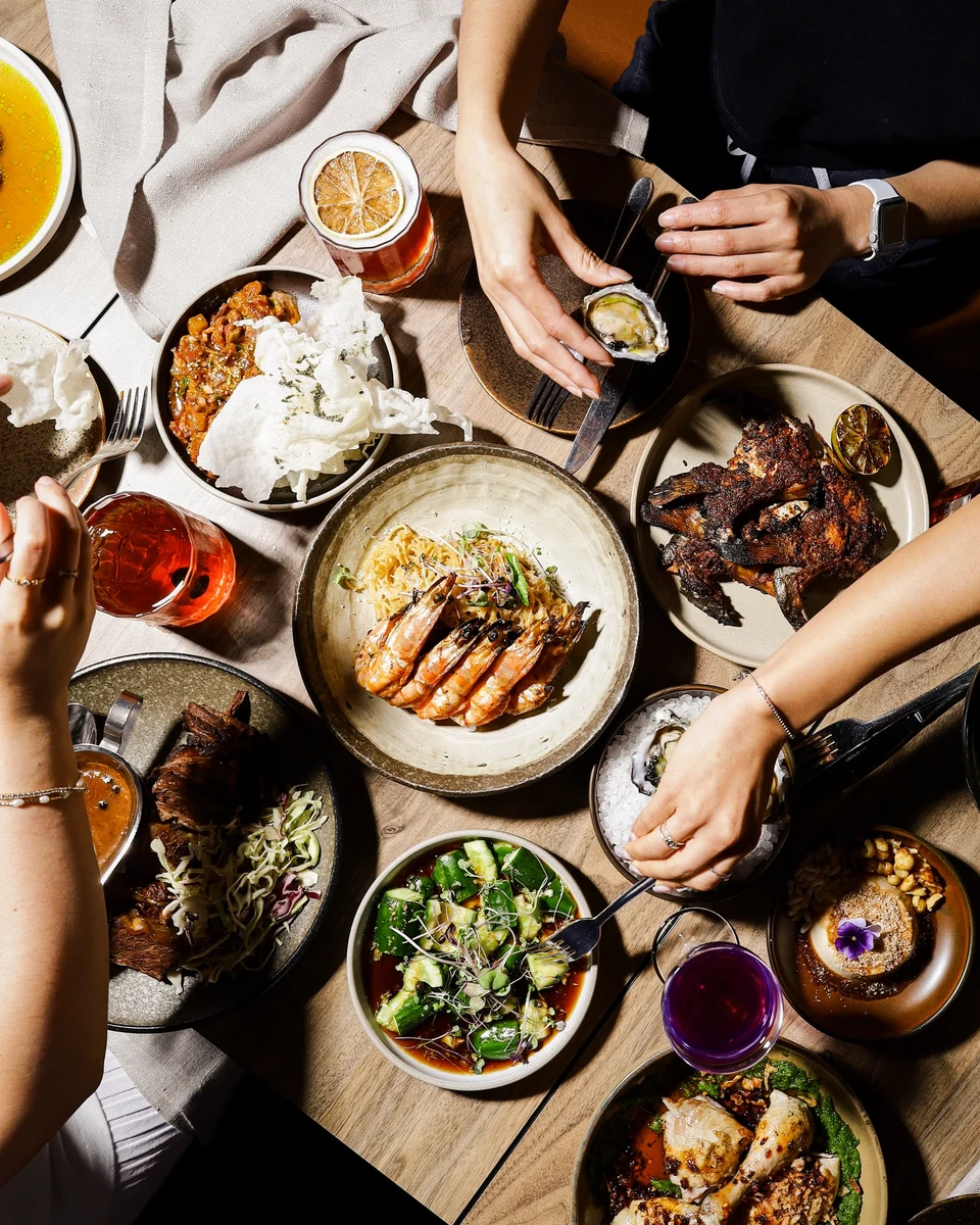 An overhead view of diners’ hands around a table of prawns, oysters, grilled food, vegetables and colourful drinks.