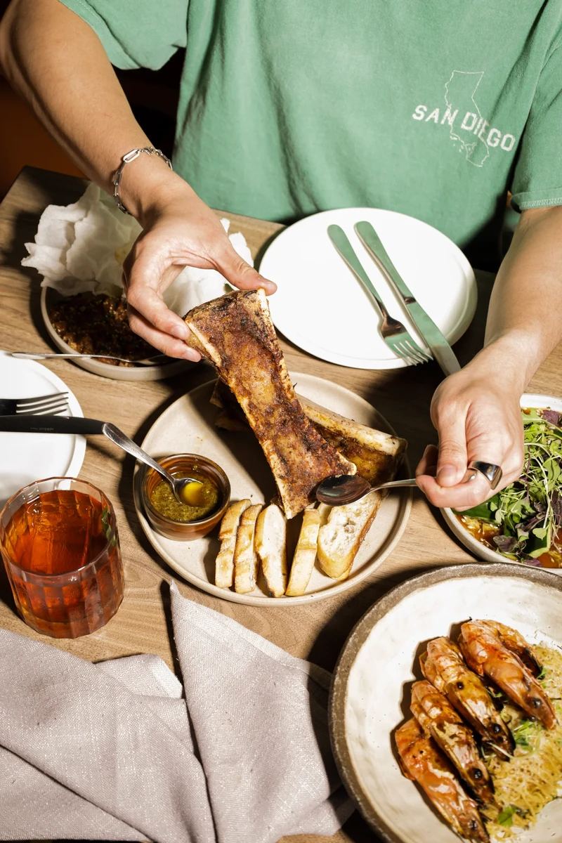 A diner holds a roasted bone and a spoon over a plate of bread on a restaurant table.