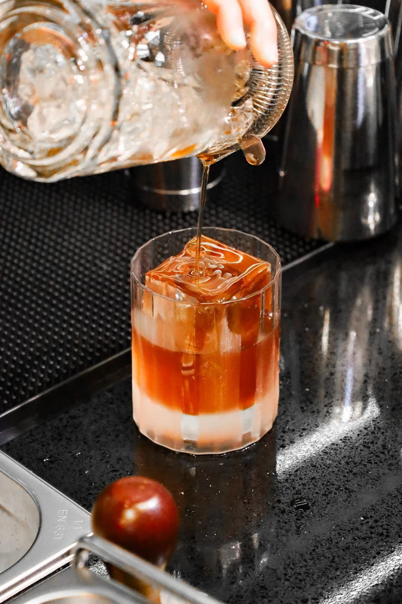 Liquid is poured from a mixing glass into a short glass of amber liquid and ice on a black bar counter.