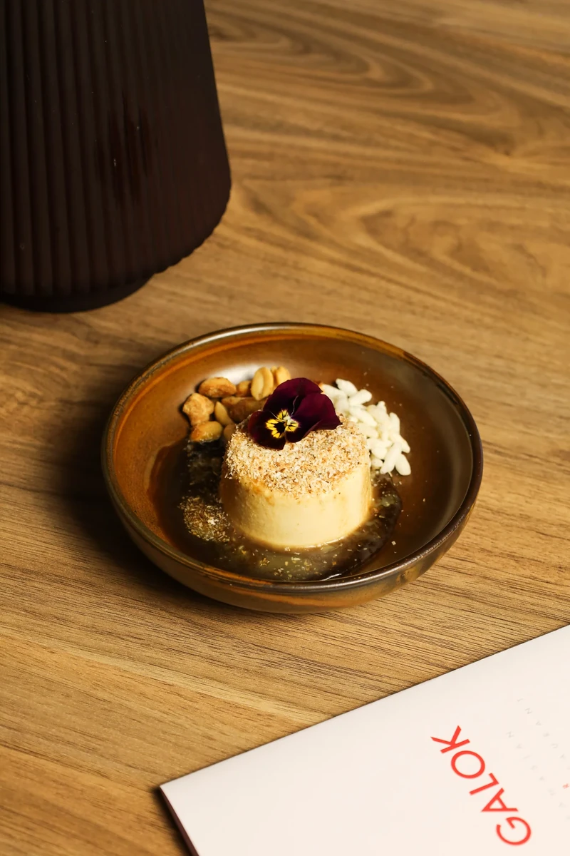 A small round dessert topped with a dark garnish sits on a brown plate beside a white menu.