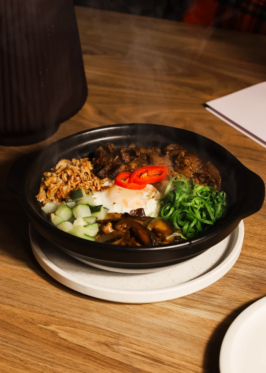 A black bowl holds rice, vegetables, sliced mushrooms and a red chilli garnish on a wooden table.