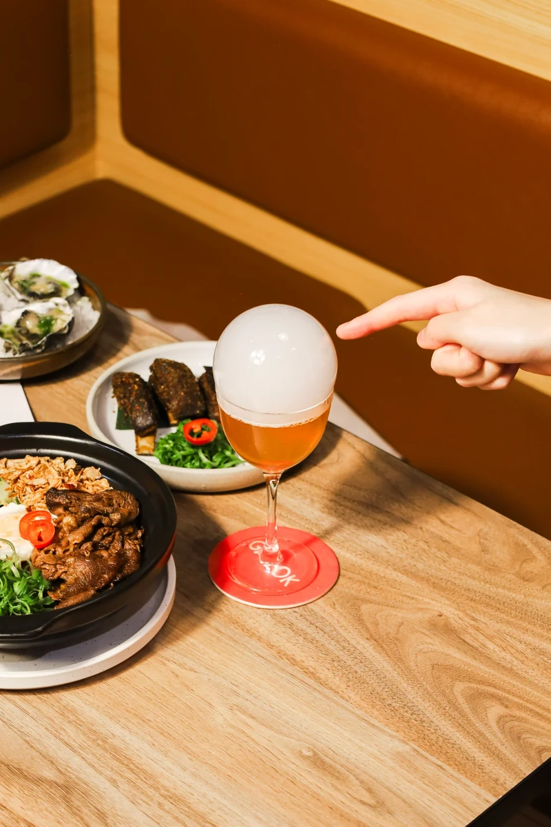 A hand points towards a pale sphere resting above a drink, with food on the table and a tan booth behind it.