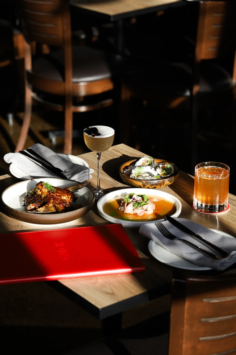 Sunlight falls across a table of food, a drink, folded napkins and a red menu in a dark dining room.