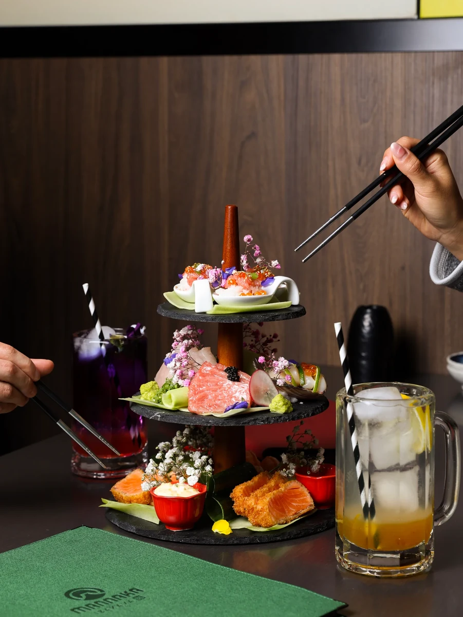 Hands hold chopsticks around a three-tier food stand, with two drinks and a green menu on the table.