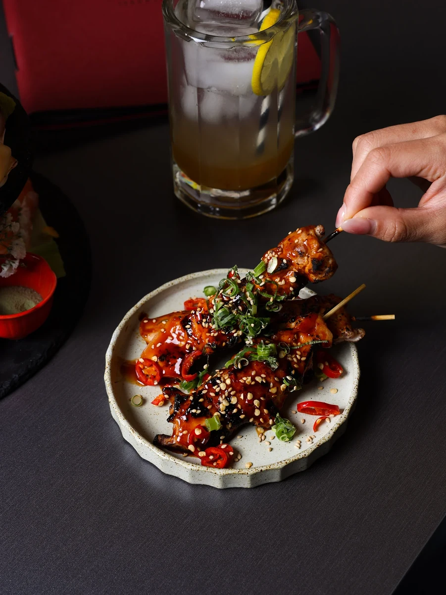 A hand reaches for a glazed skewer on a small plate beside an iced drink.
