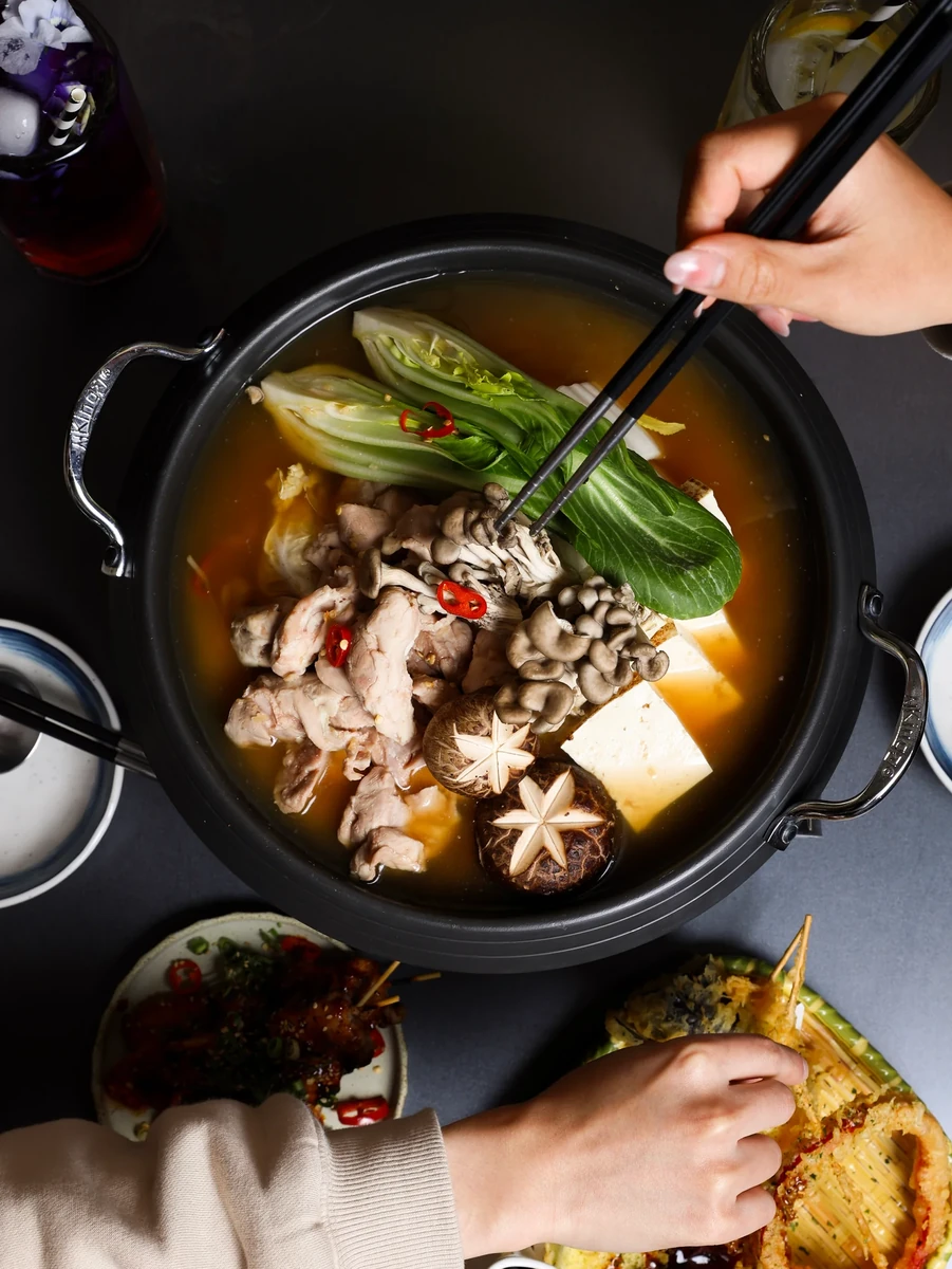 A hand holds a spoon above a black pot containing sliced meat, mushrooms and vegetables.