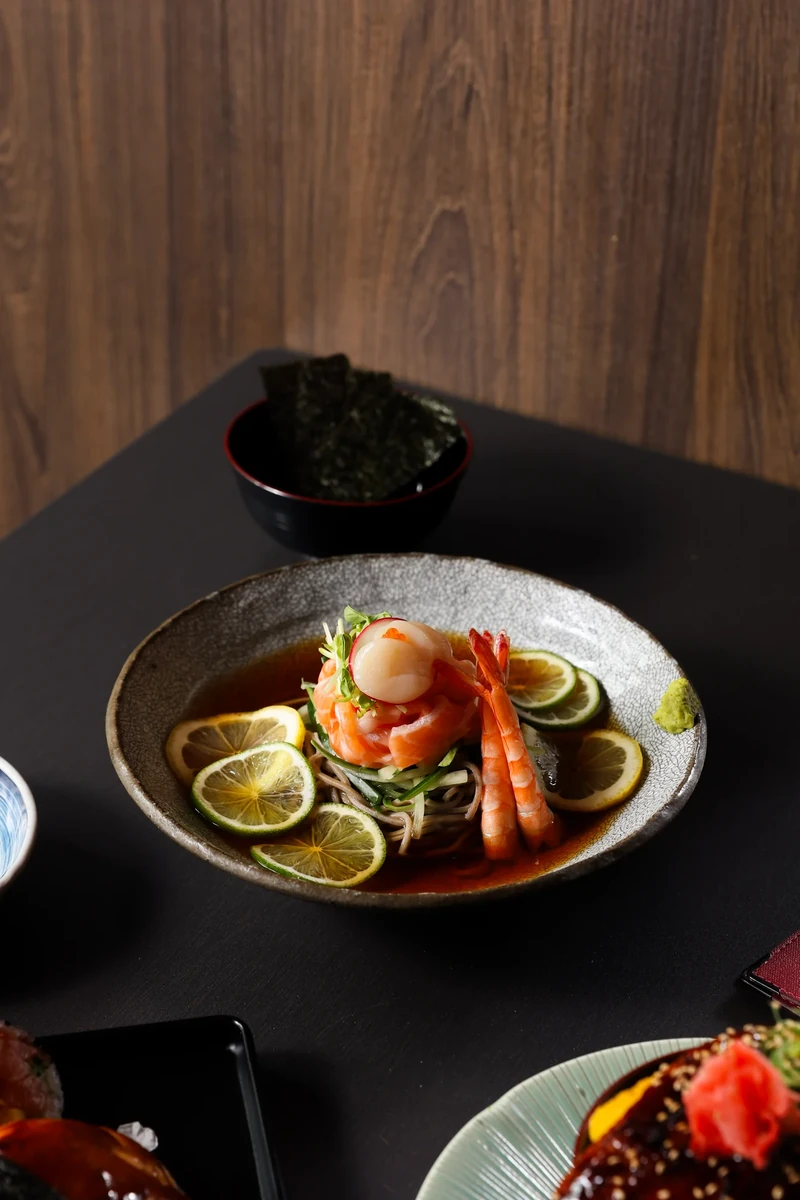 A grey plate of seafood and thin citrus slices sits on a dark table in front of a timber wall.