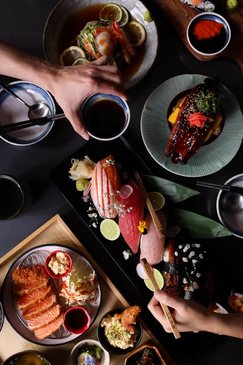 An overhead view of sliced fish, glazed fish, breaded salmon and other dishes, with hands holding a cup and chopsticks.