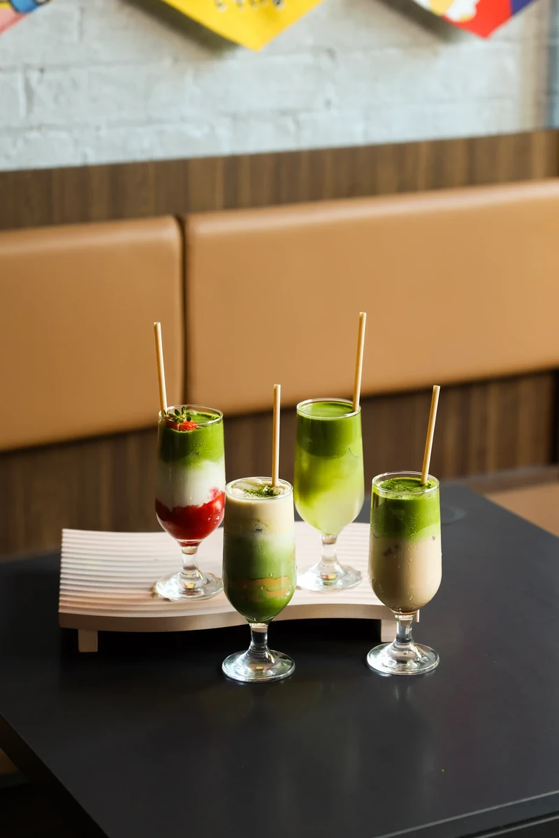 Four green drinks in tall glasses stand on a dark table in front of a tan upholstered booth.