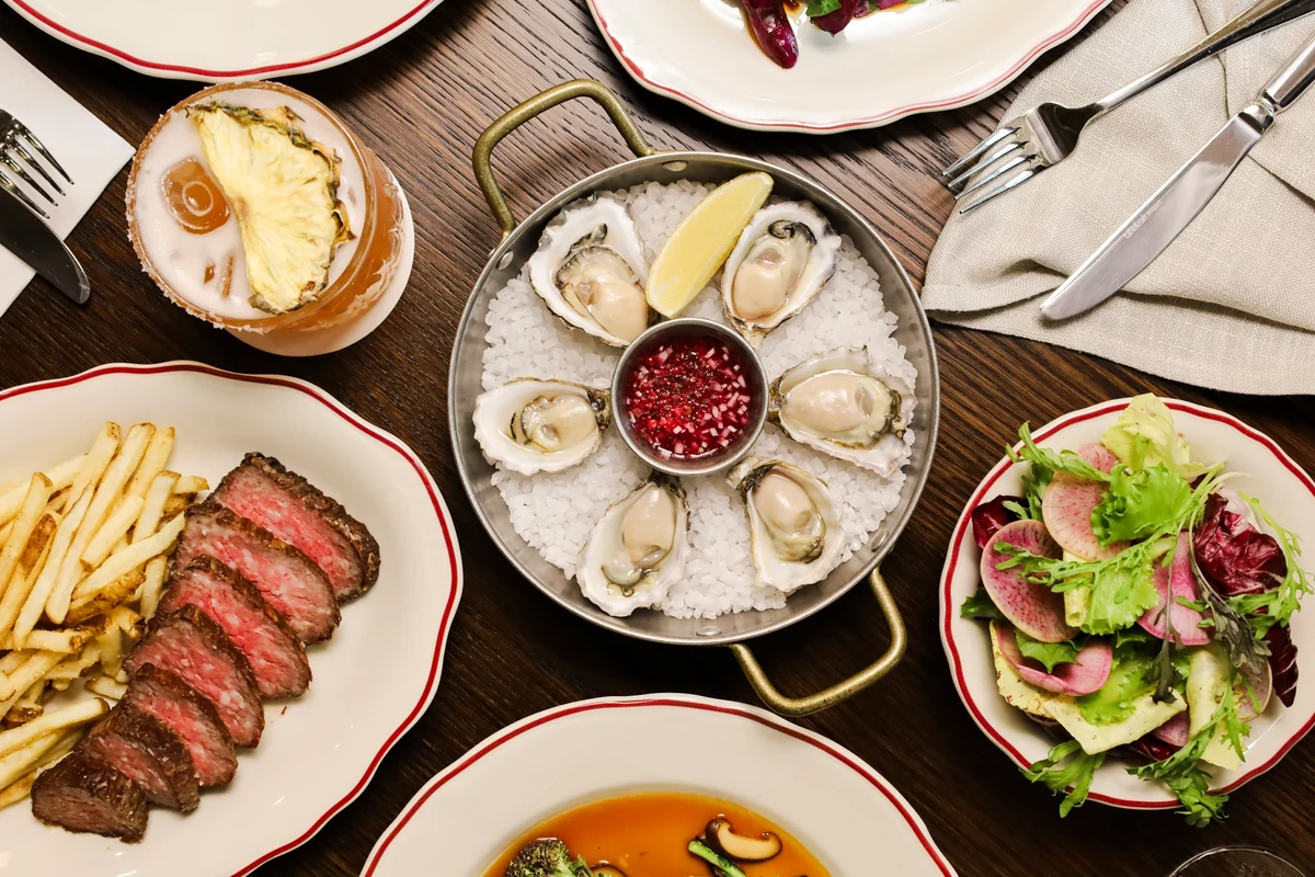 An overhead view of oysters, sliced steak, chips, salad and drinks on a wooden table with white napkins.