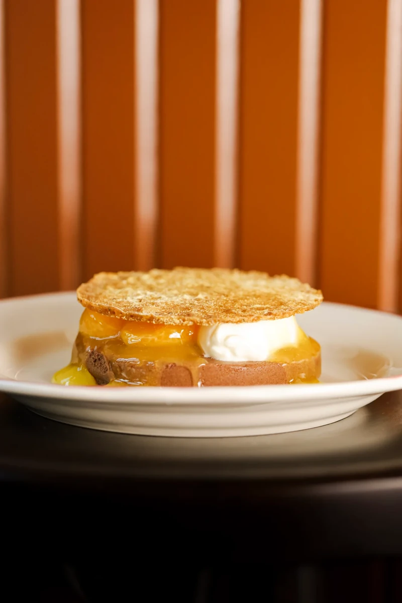 A layered dessert with cream between golden discs sits on a white plate against an orange and cream background.