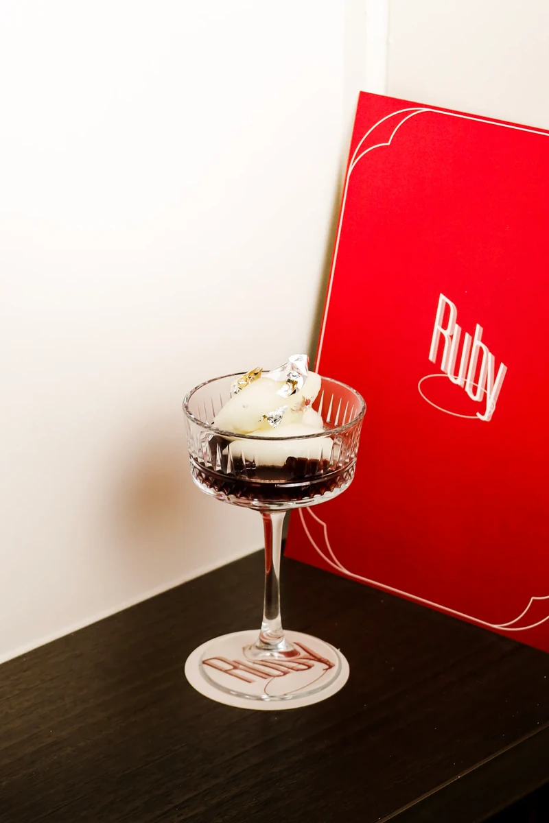 A stemmed glass containing a dark dessert topped with pale scoops stands in front of a red menu.
