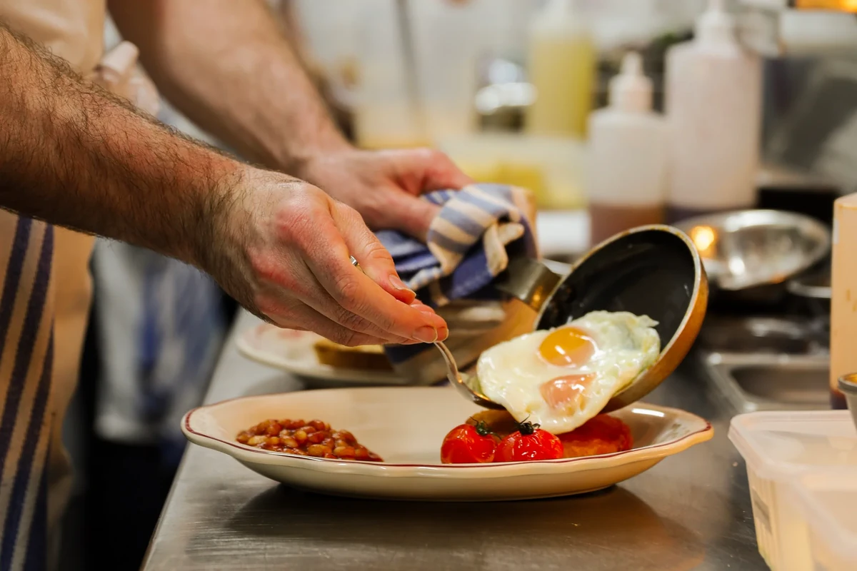 Hands slide a fried egg from a pan onto a breakfast plate with tomatoes.