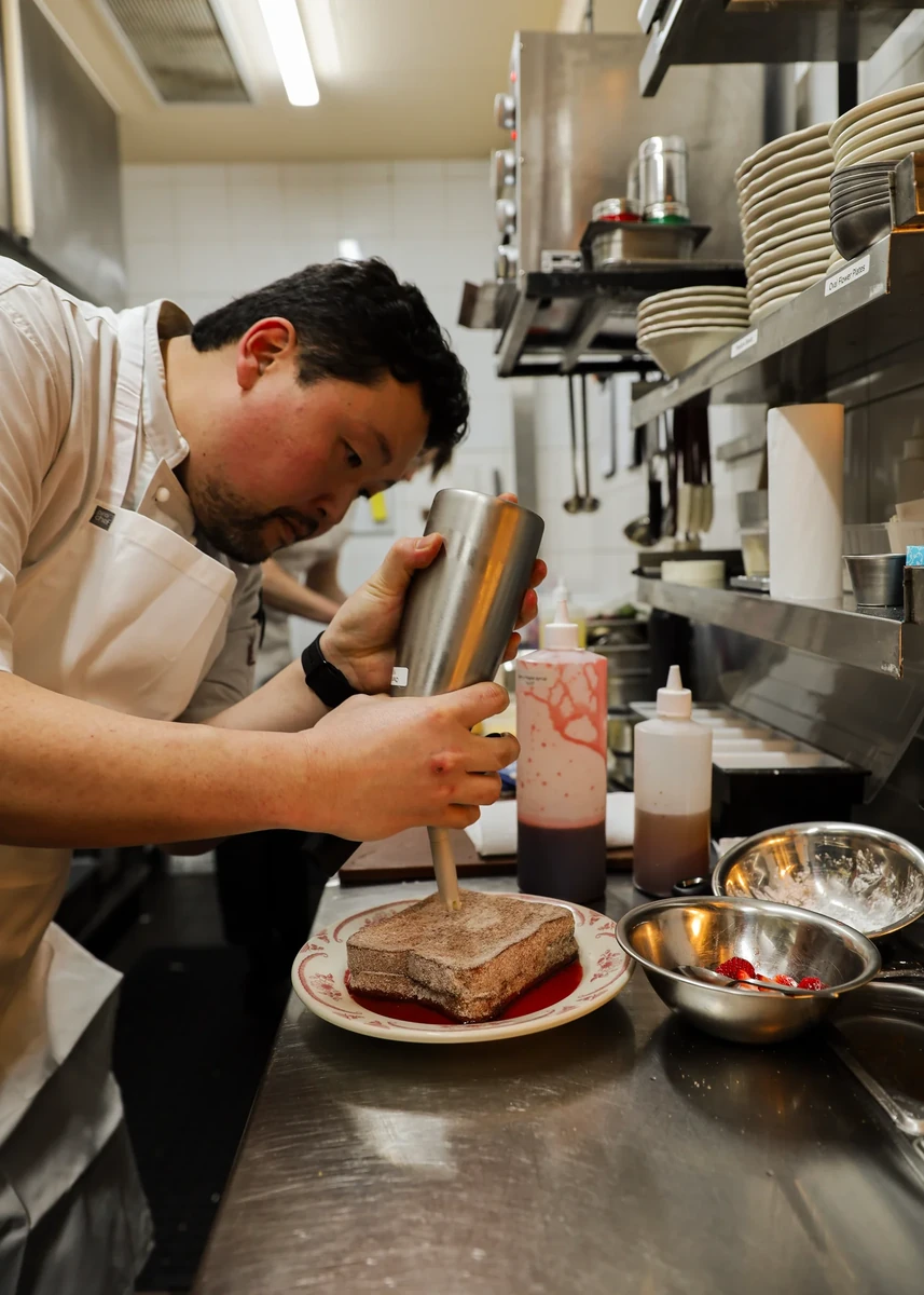 A person in an apron dispenses a topping onto thick toast in a stainless-steel kitchen.