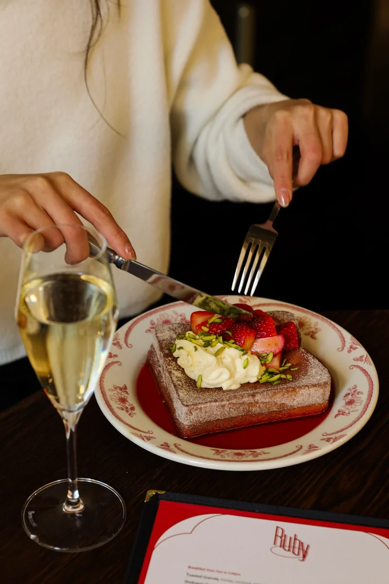 A diner cuts into thick toast topped with cream, strawberries and chopped green nuts, beside a pale drink and a red menu.