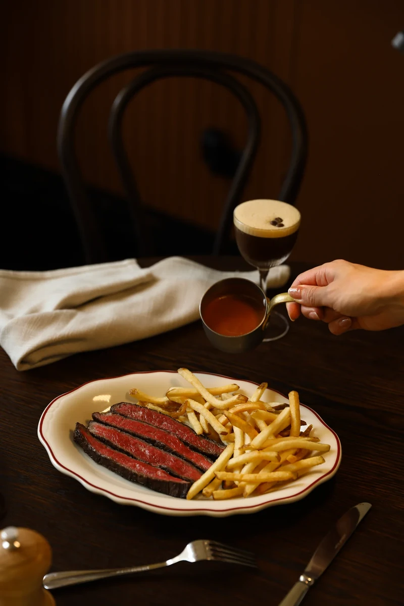 A hand holds a small stemmed drink beside a plate of sliced steak and chips on a dark wooden table.