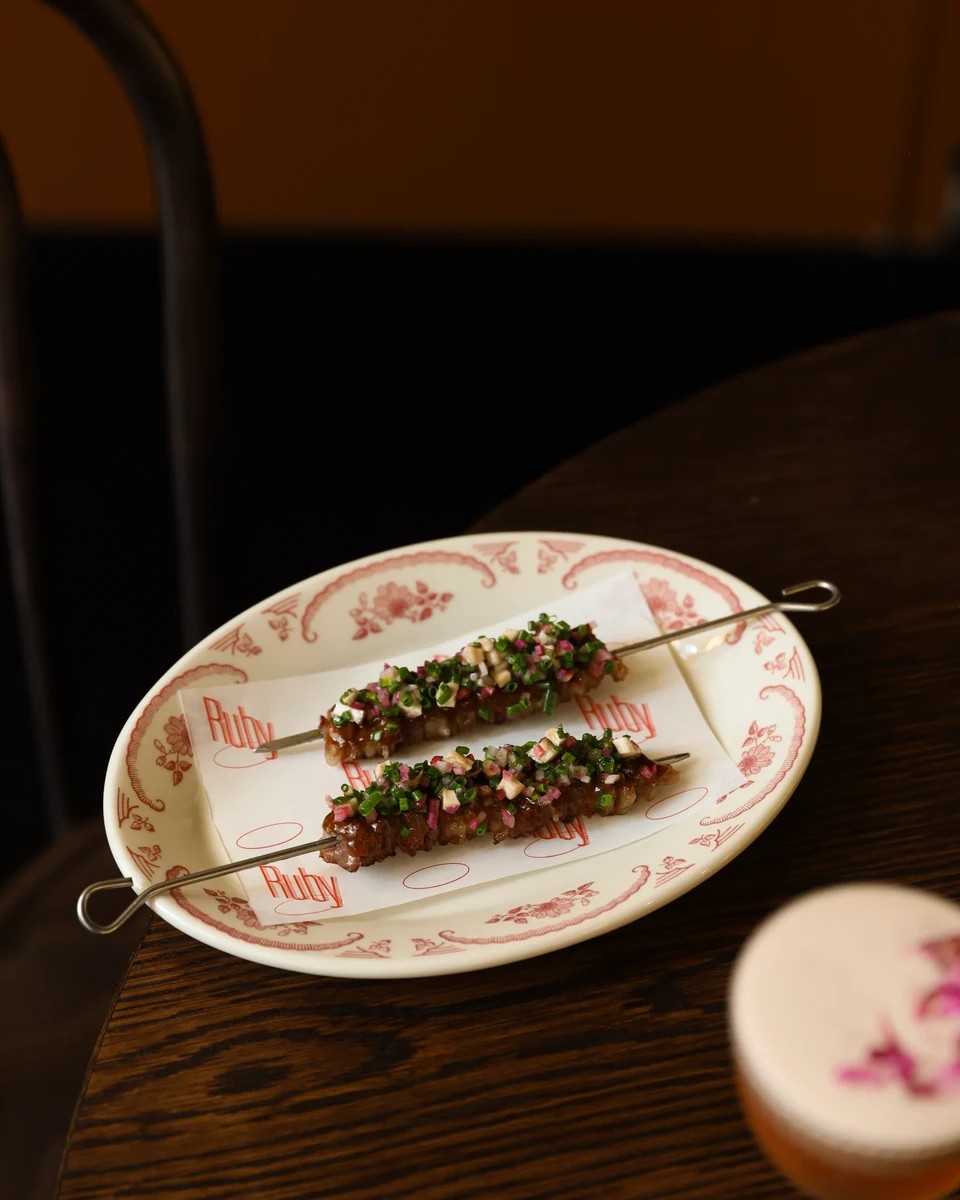 Two garnished skewers rest on a white plate with a red floral border on a dark wooden table.