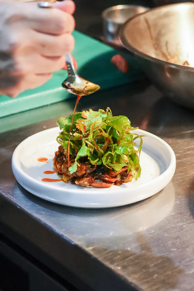 A hand spoons sauce over a plated dish on a metal kitchen counter.