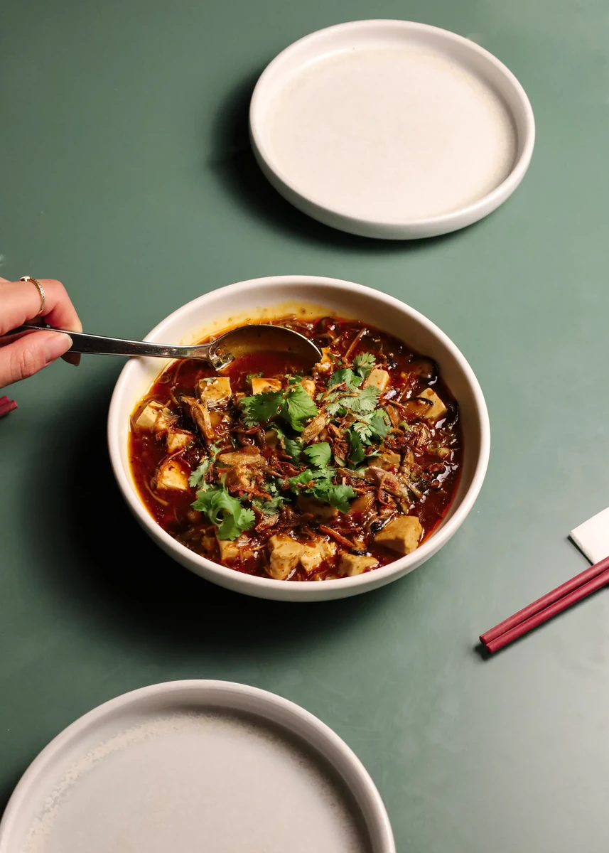 A hand holds a spoon over a bowl of food on a green table.