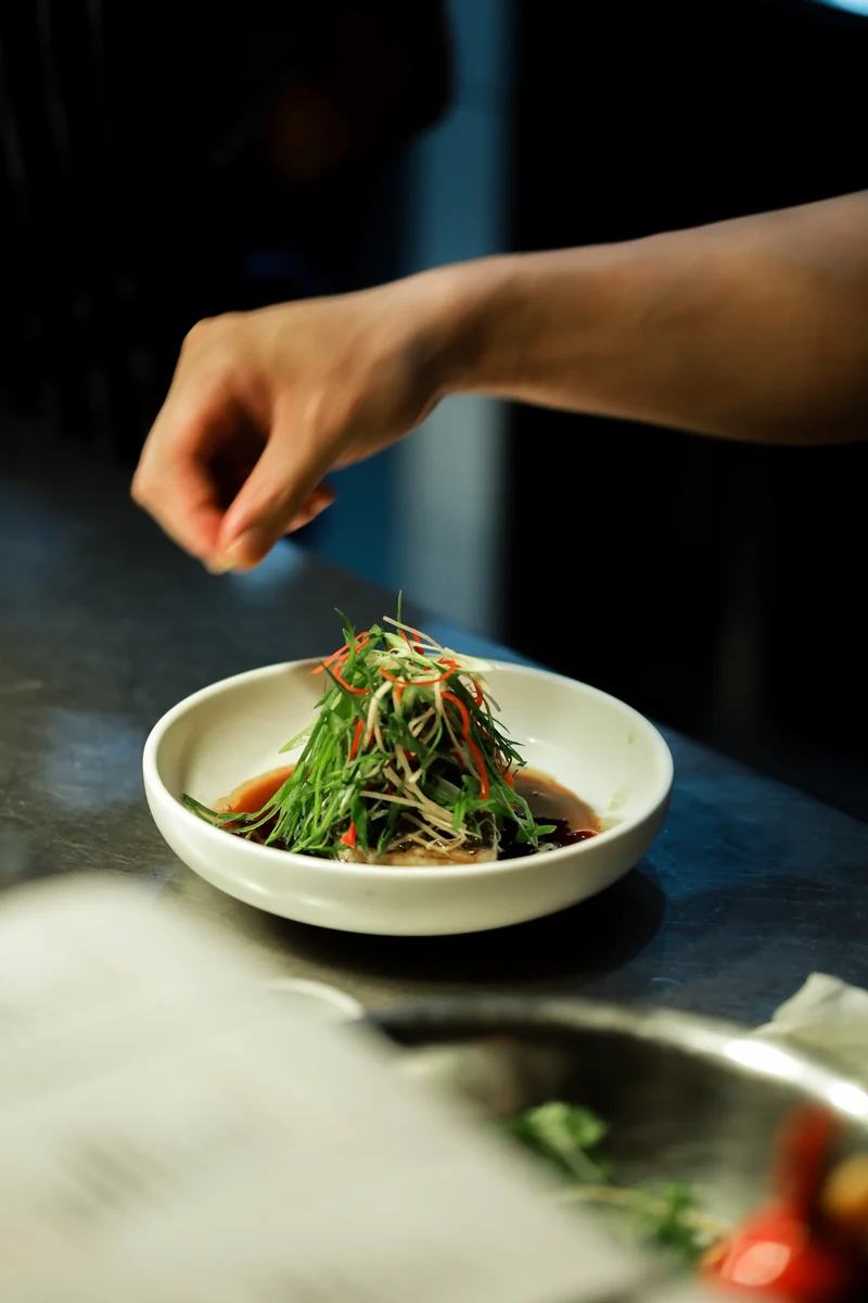 A hand garnishes a bowl of food in a shaft of sunlight.
