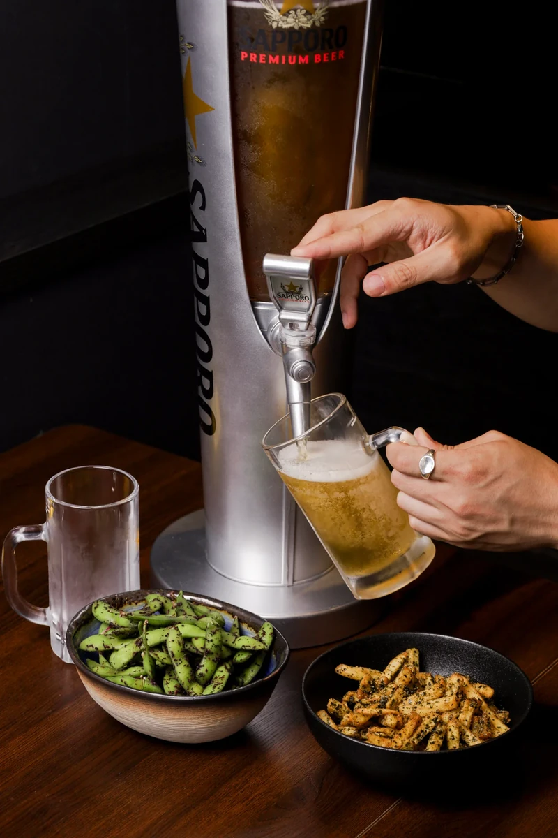 Hands pour beer from a tall dispenser into a mug beside small dishes.