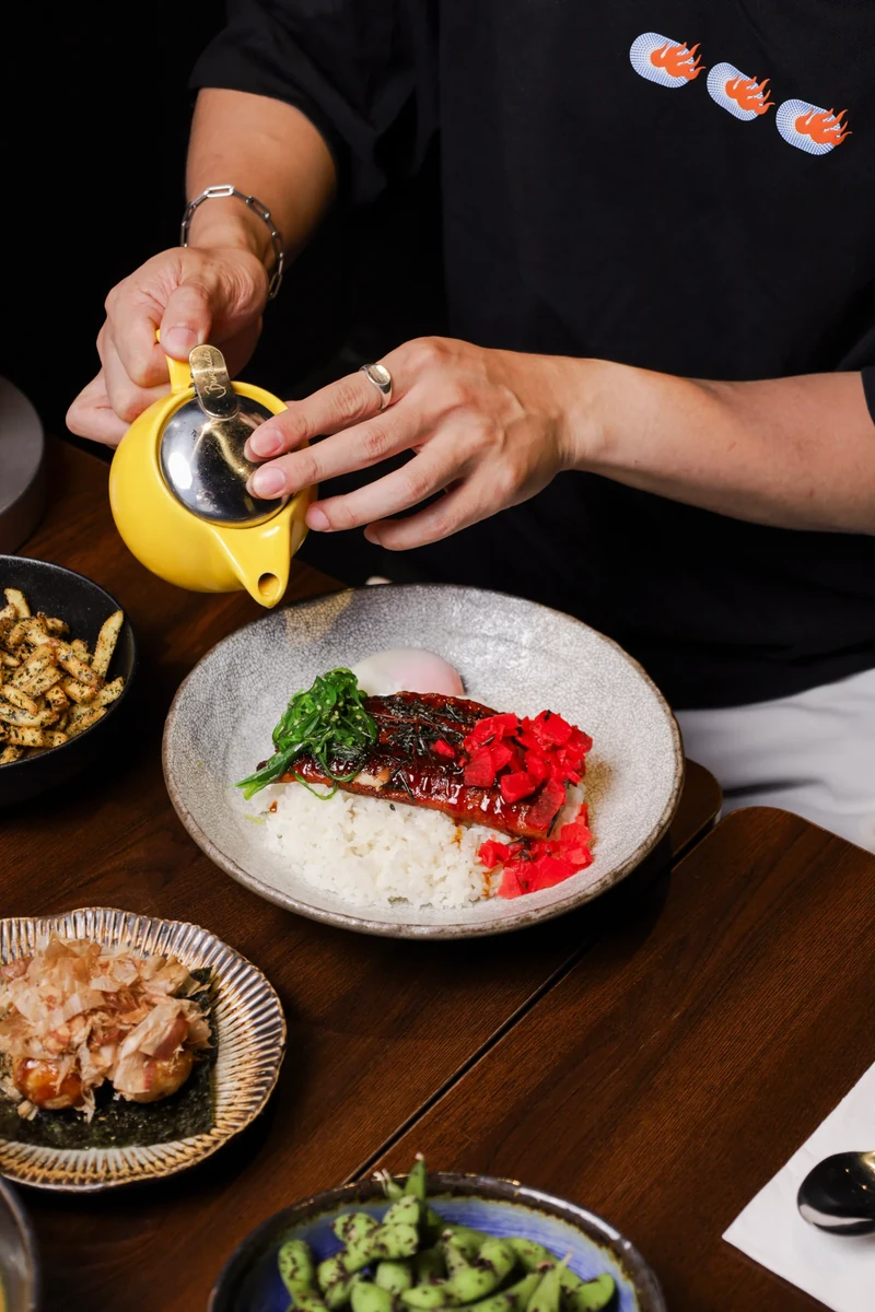 Hands pour from a small yellow pot over a bowl of rice with red and green toppings.