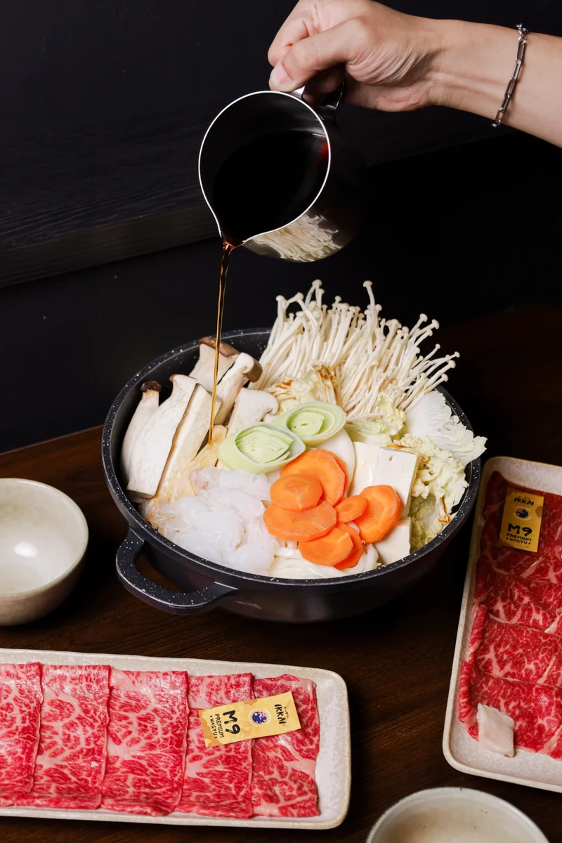 A hand pours broth into a pot filled with vegetables beside a tray of sliced beef.