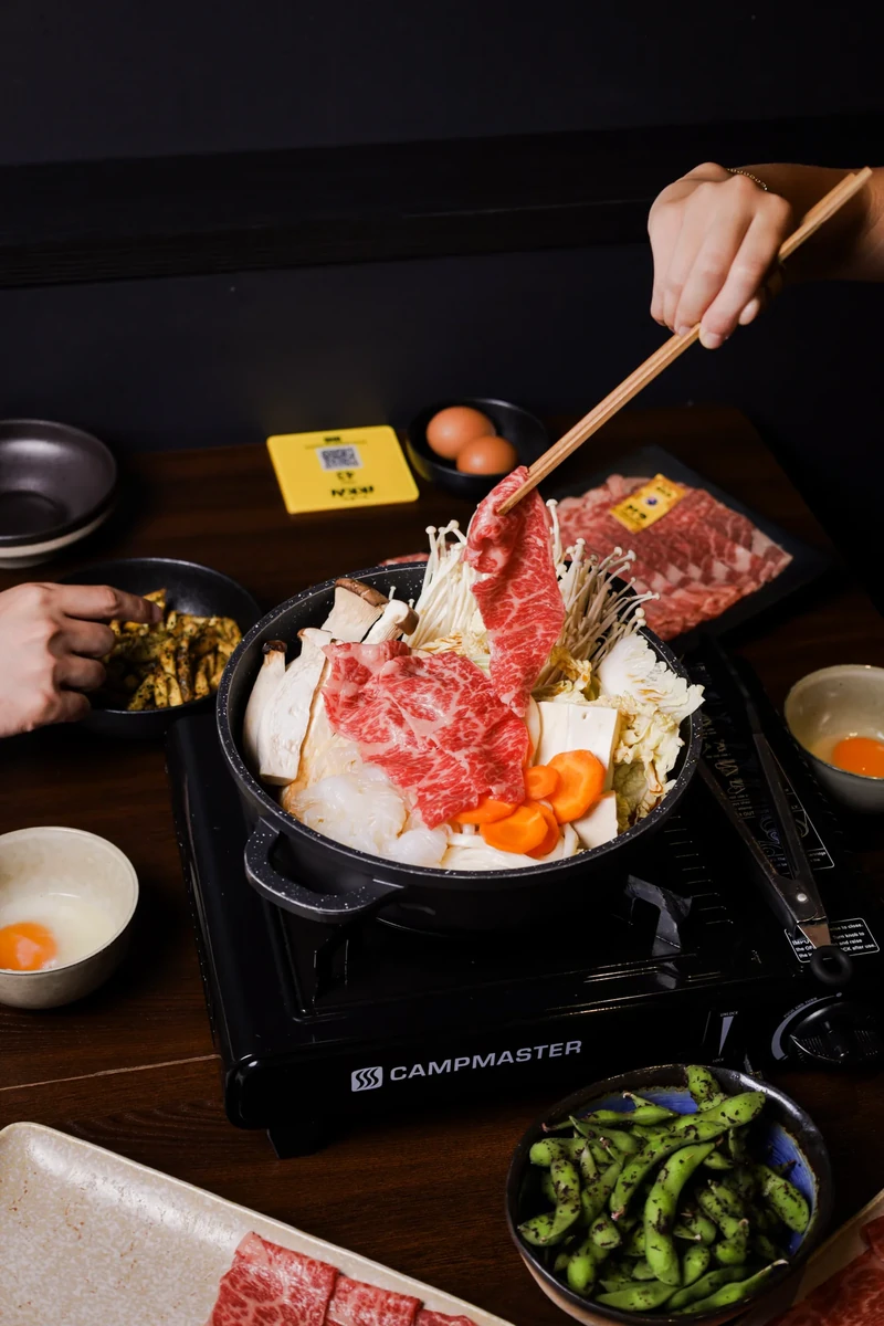 Chopsticks lift a slice of beef above a hotpot of vegetables on a table.