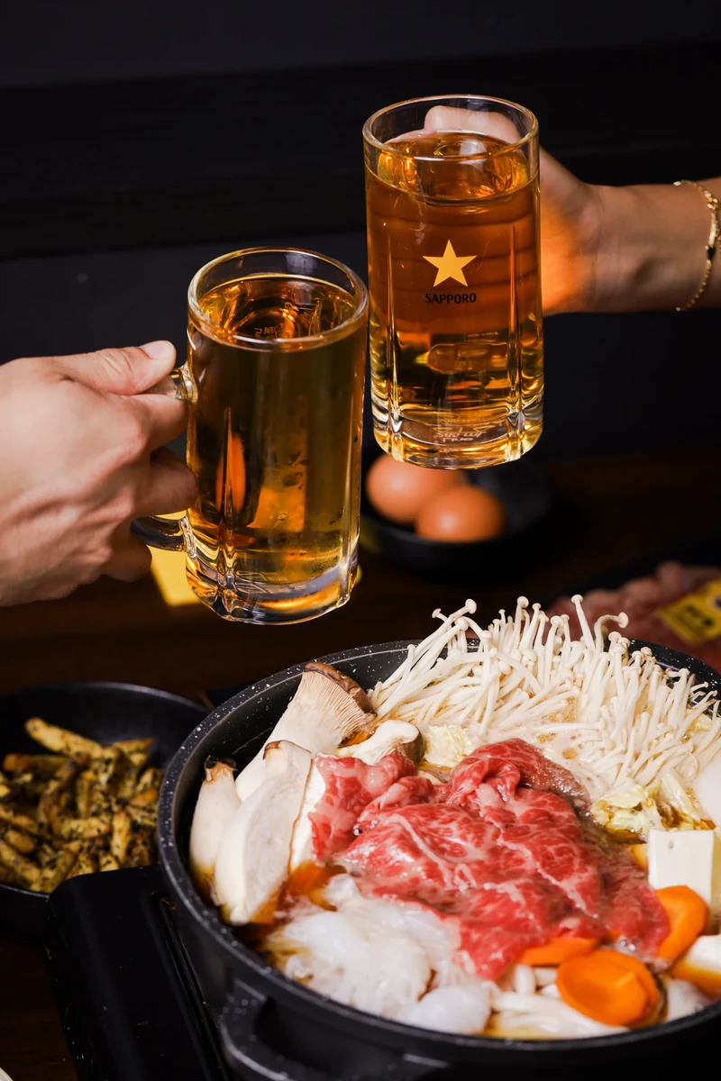Two hands raise beer mugs above a hotpot on the table.