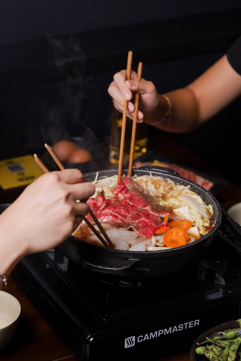 Hands with chopsticks reach into a steaming hotpot.