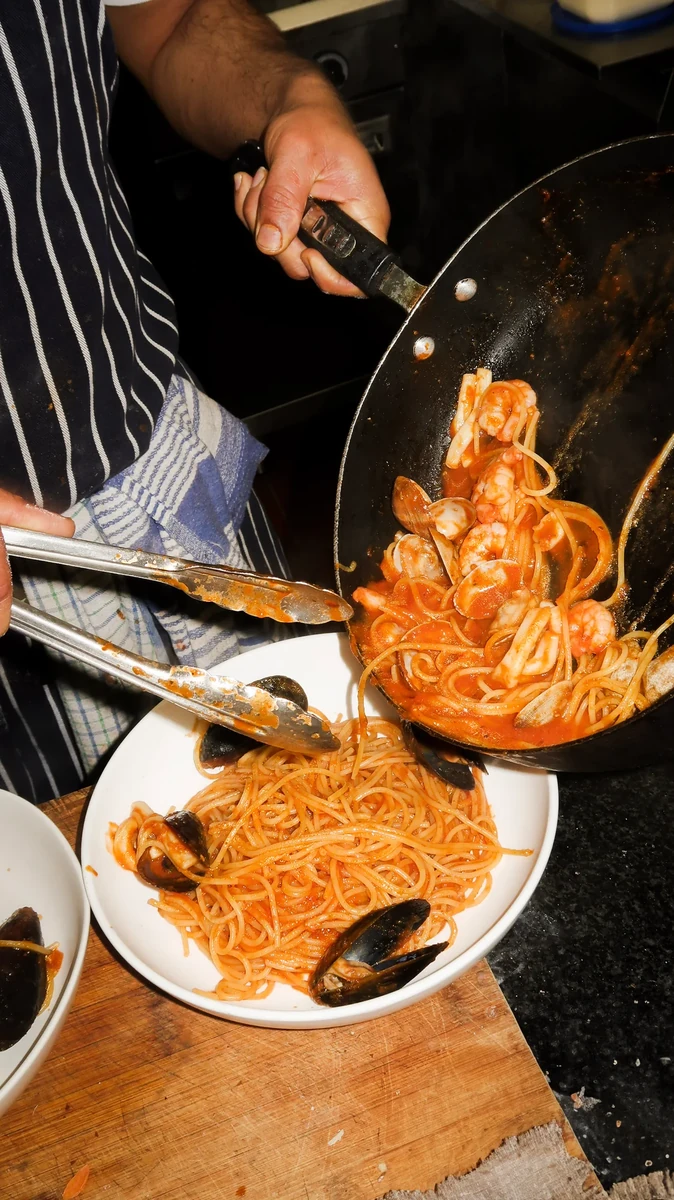 A person tips tomato-coated pasta and mussels from a pan into a white plate.