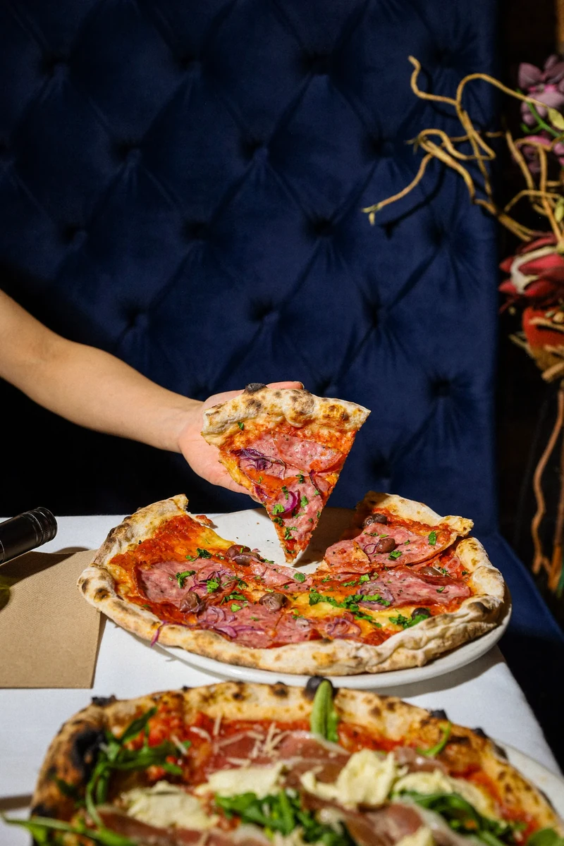 A hand lifts a pizza slice above a pizza on a white tablecloth in front of a blue upholstered seat.