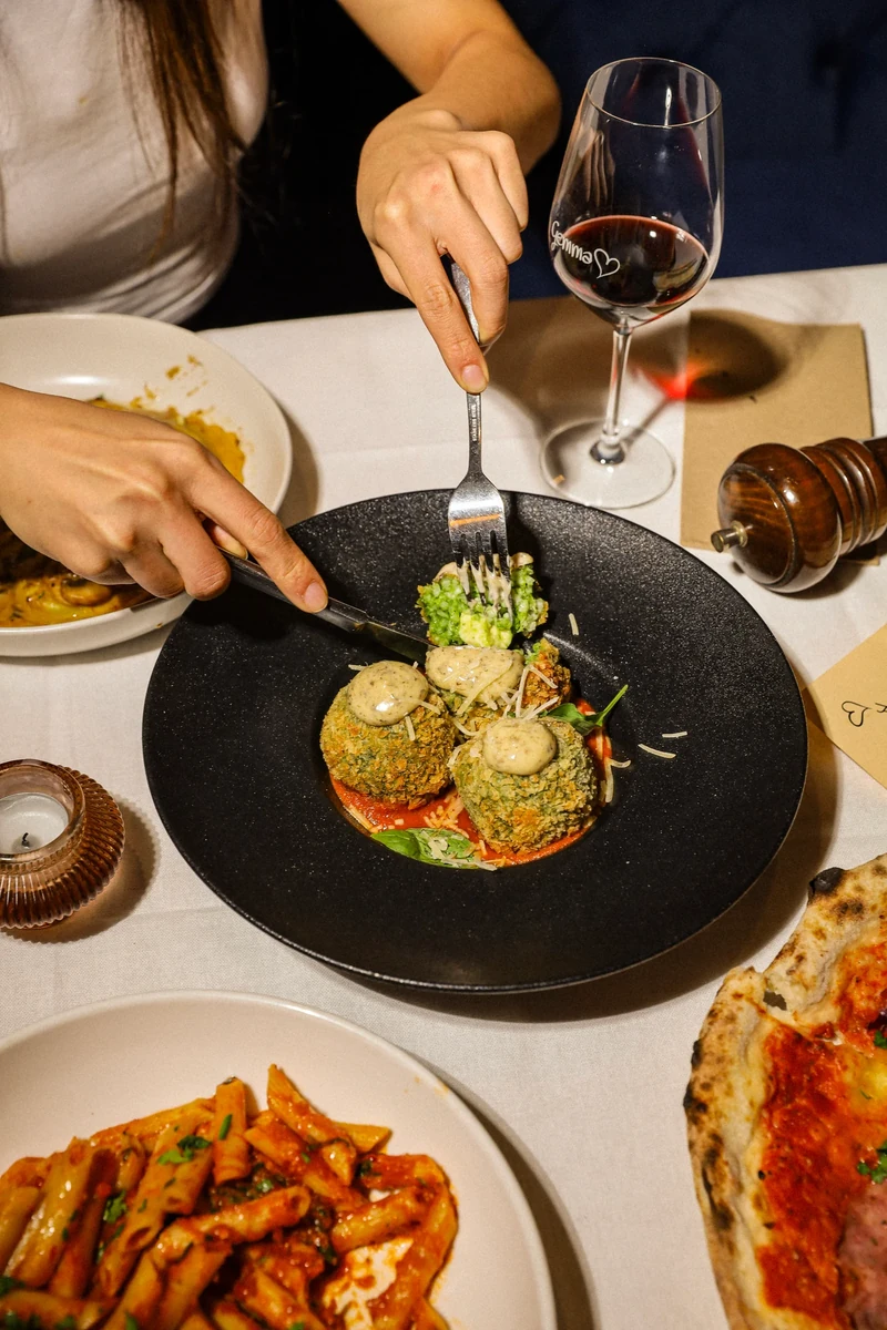 A diner uses a fork and knife over a black plate of round pieces with greens, beside a glass of red wine.