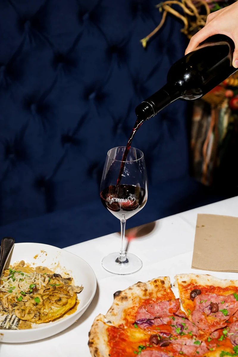 Red wine is poured into a wine glass, with pizza and pasta on the table below.