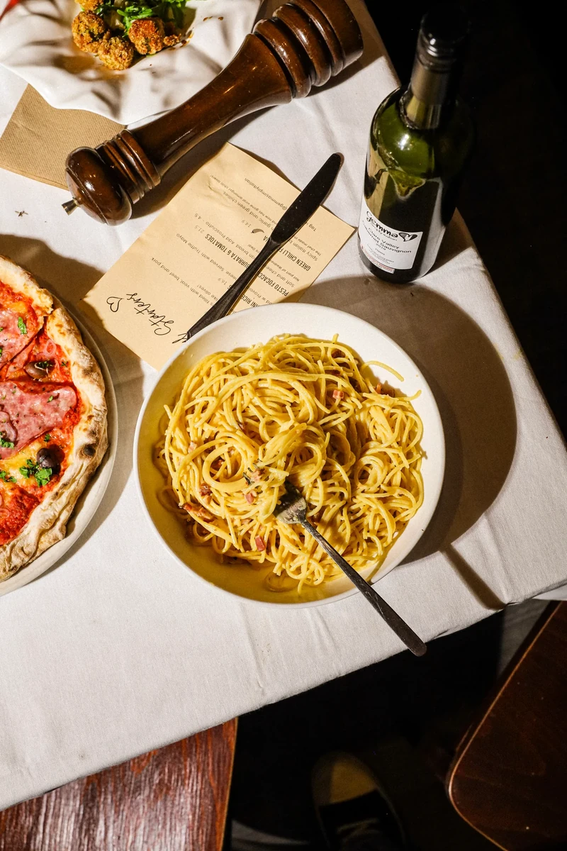 A bowl of yellow pasta sits on a white tablecloth beside a wine bottle, pepper mill and menu.