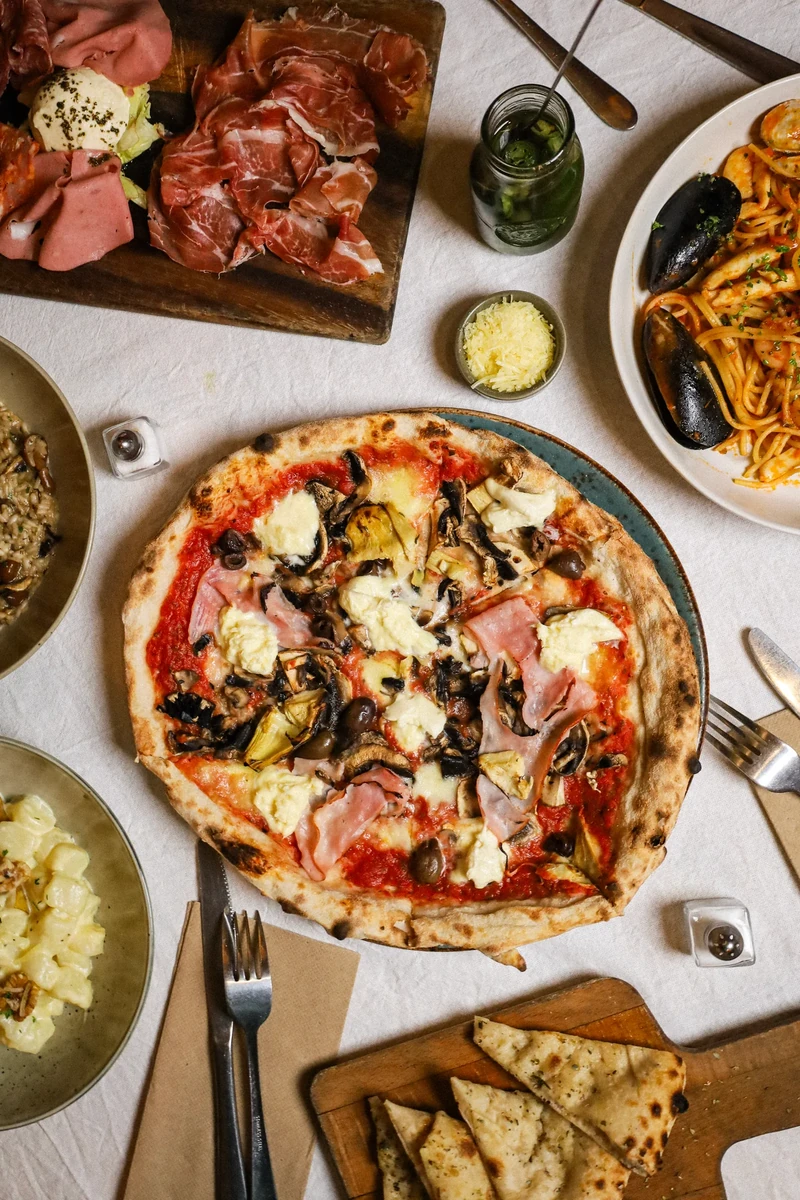 An overhead view of a pizza on a white tablecloth, surrounded by sliced meats, bread and other dishes.