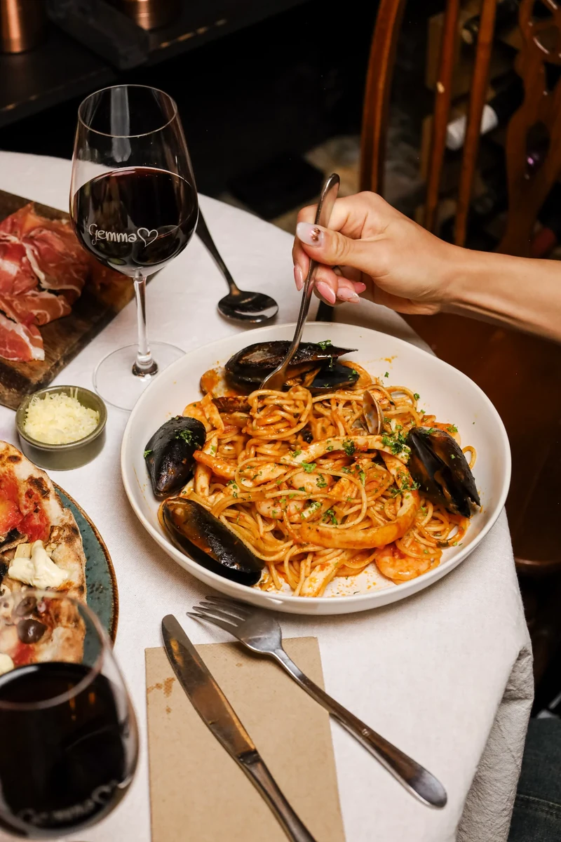 A hand holds a fork over a bowl of tomato-coated pasta and mussels beside a glass of red wine.