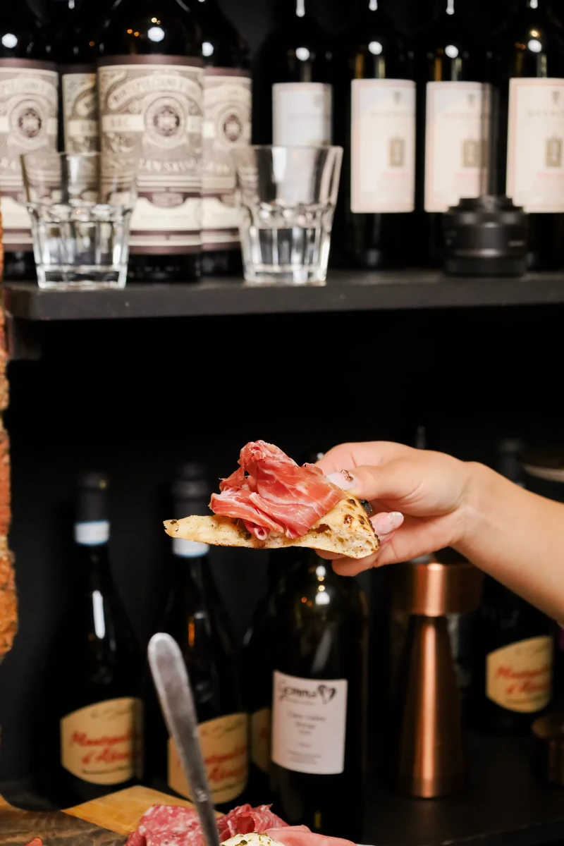 A hand holds bread topped with folded slices of cured meat in front of shelves of bottles.